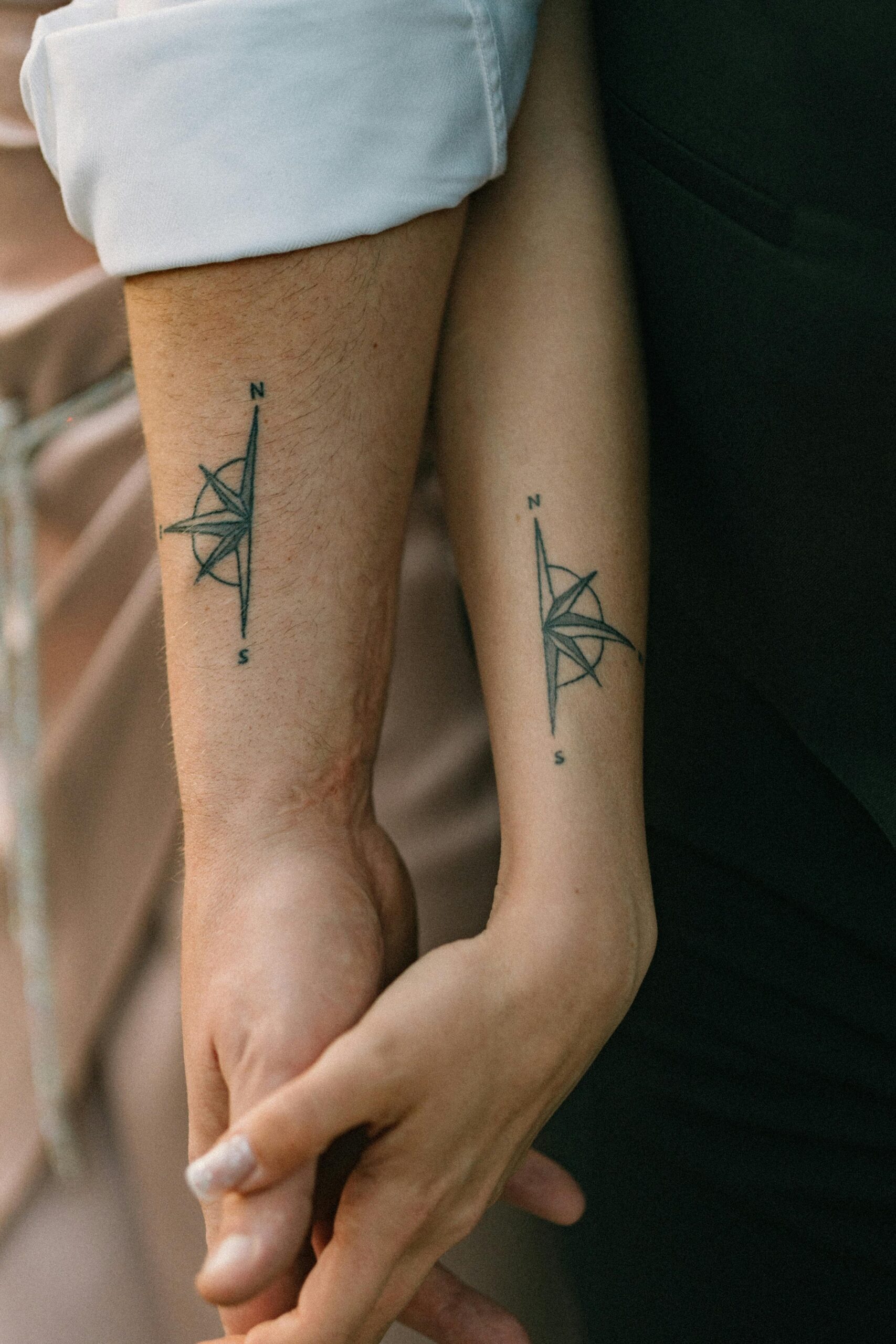 Close-up of a couple holding hands, showcasing matching compass tattoos on their forearms.